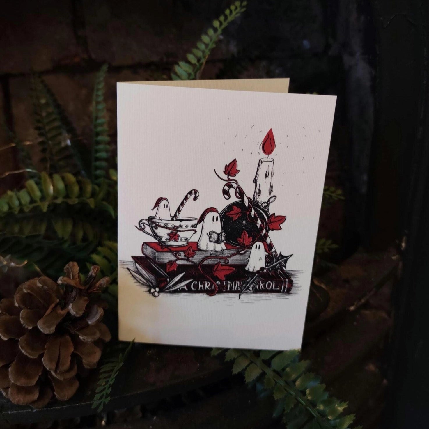 White Christmas card with nightcapped ghosts sat on a copy of A Christmas Carol, by candleight. Card is surrounded by foliage and pine cones.