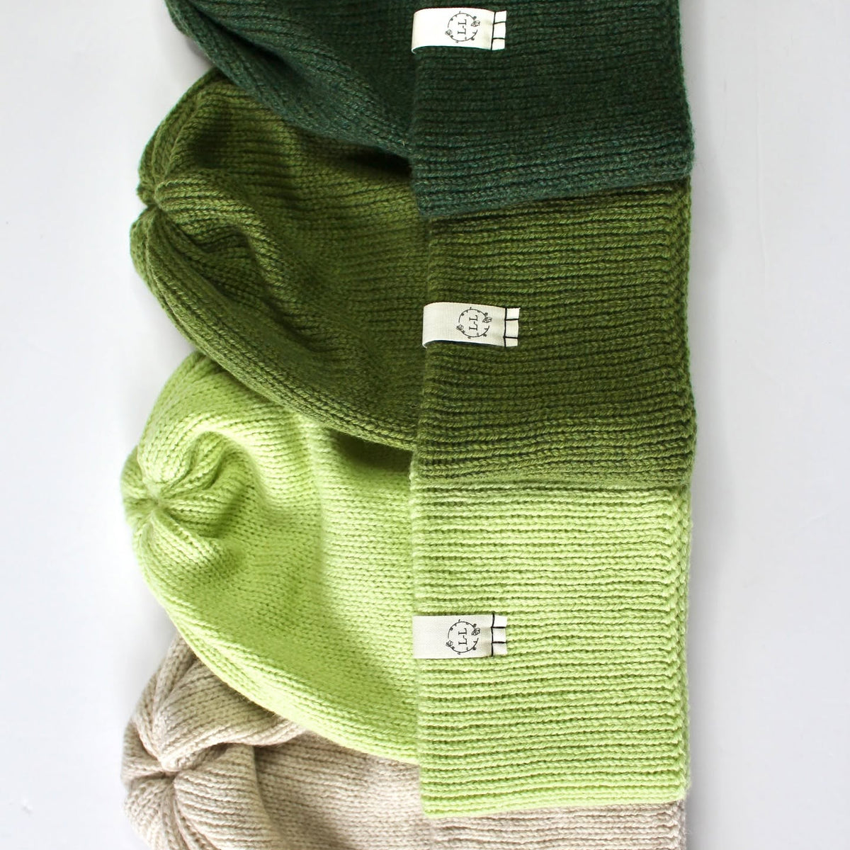 Four knitted beanie hats stacked vertically, each paired with a matching ribbed cuff that fades from a darker shade to a lighter shade. The colors shown are dark forest green, olive green, light lime green, and beige.