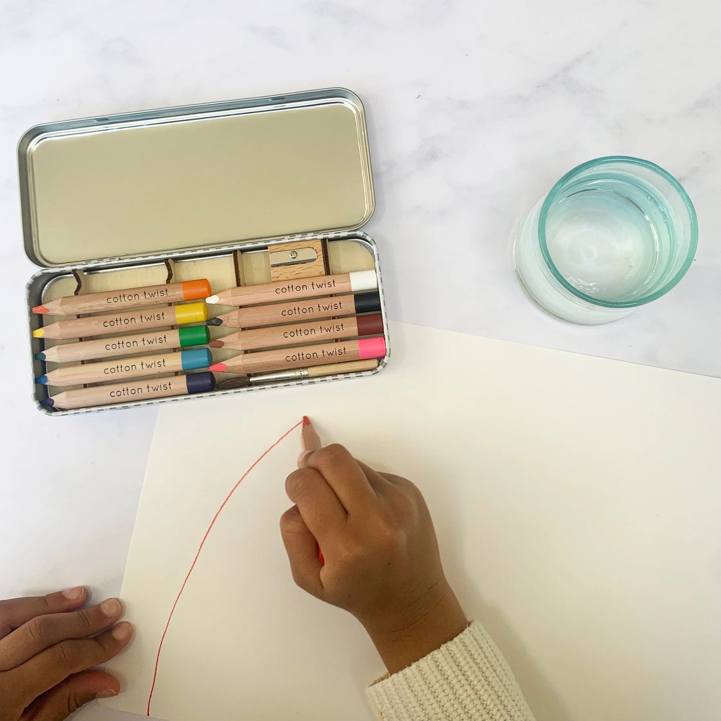 Person using colorful watercolour pencils with a tin on a white surface