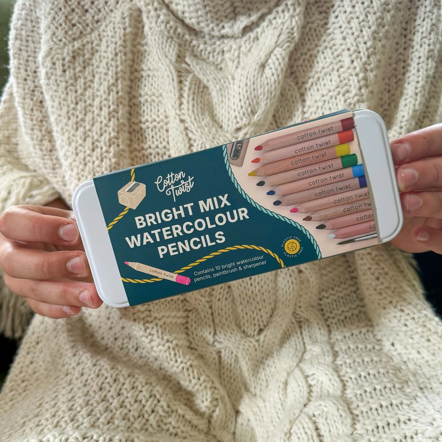 Person holding a set of Cotton Twist Bright Mix Watercolour Pencils against a textured beige background.