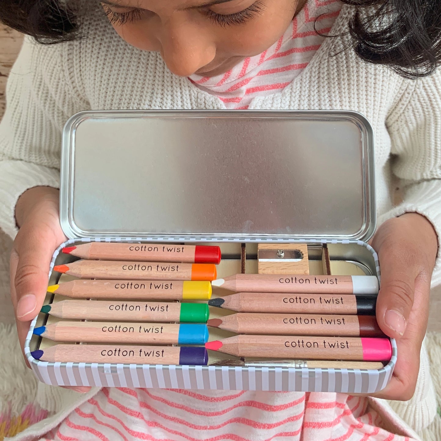 Child holding a set of Cotton Twist colored pencils in a metal tin.