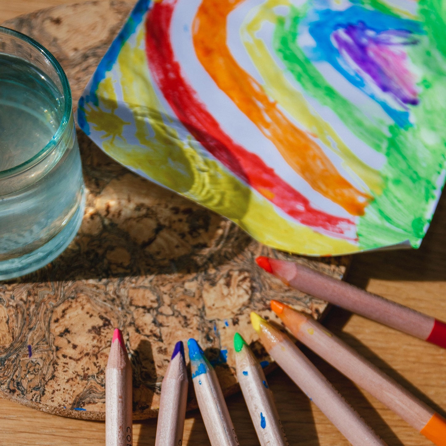 Colorful crayons and a glass of water on a wooden surface with a rainbow-colored paper.