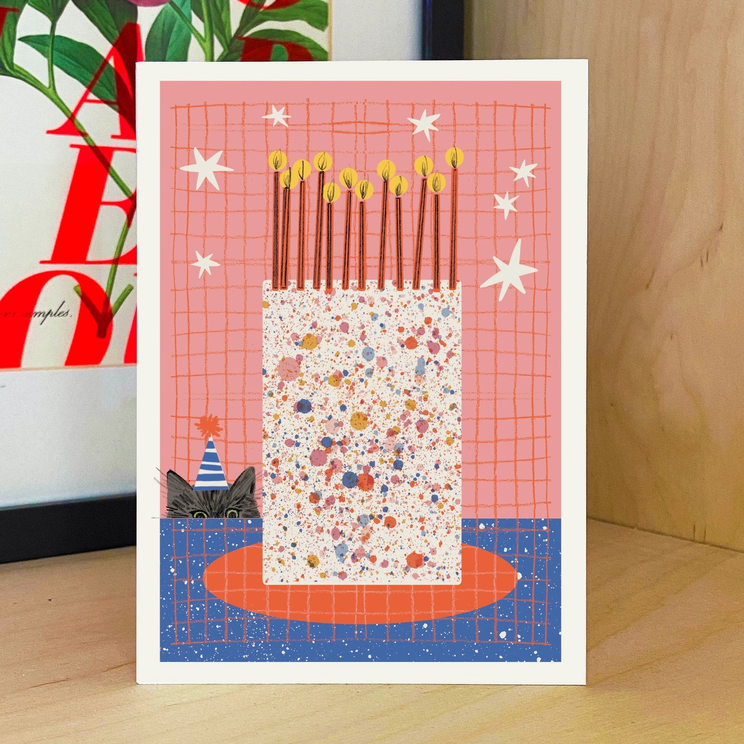 Greeting card with a cat peeking out from behind a big colourful birthday cake with candles, on a wooden surface.