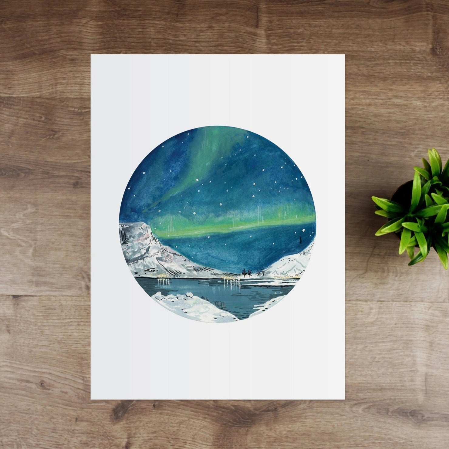 Art print of a circular design with a blue sky and green aurora on a wooden surface with a small plant.