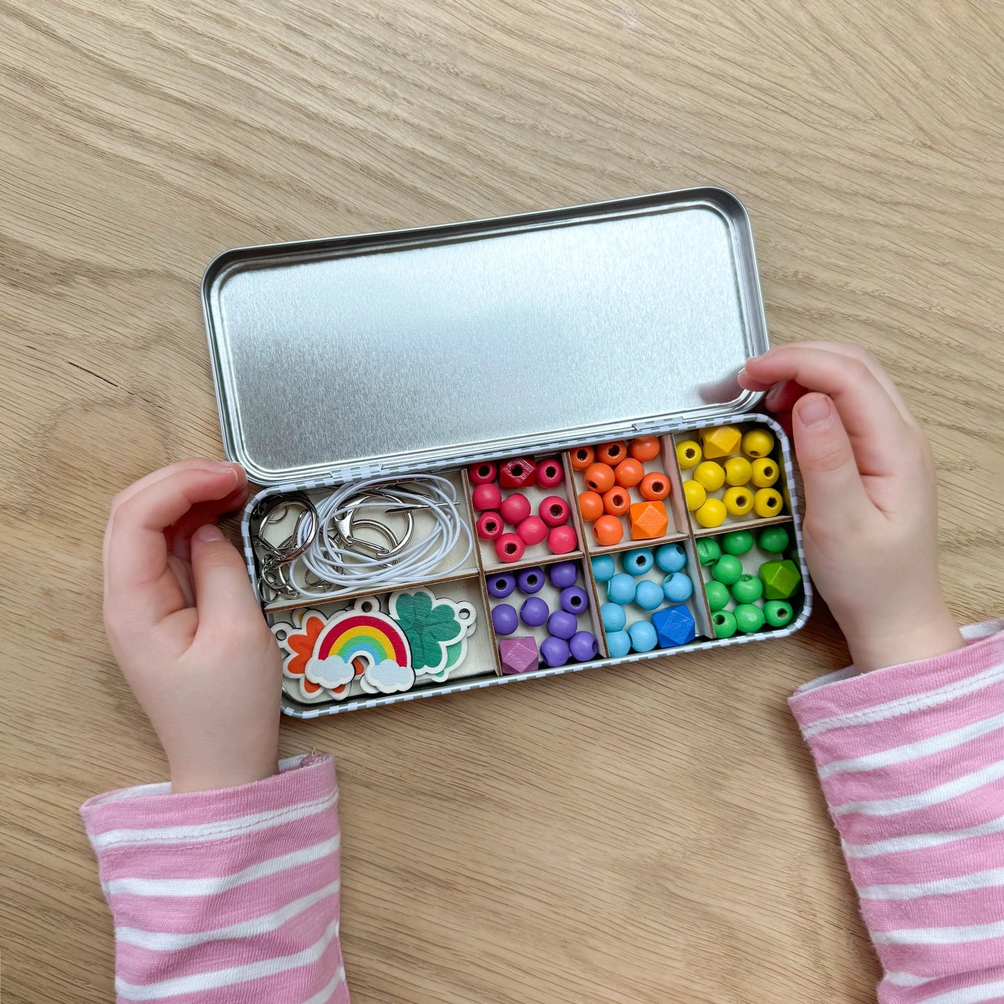 Child's hands holding a small metal box with compartments filled with colorful beads and jewelry-making supplies on a wooden surface.