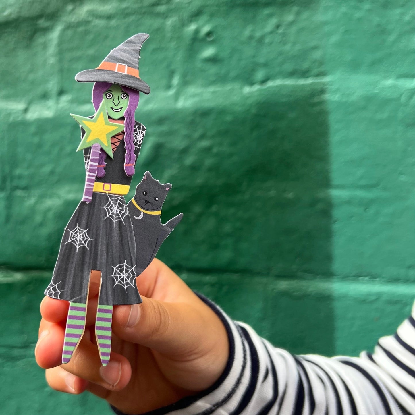 Hand holding a paper craft of a witch with a black cat against a green background