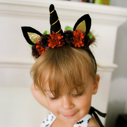 Child wearing a Halloween-themed headband with a black horn, ears and colorful flowers, sitting in front of a white fireplace.
