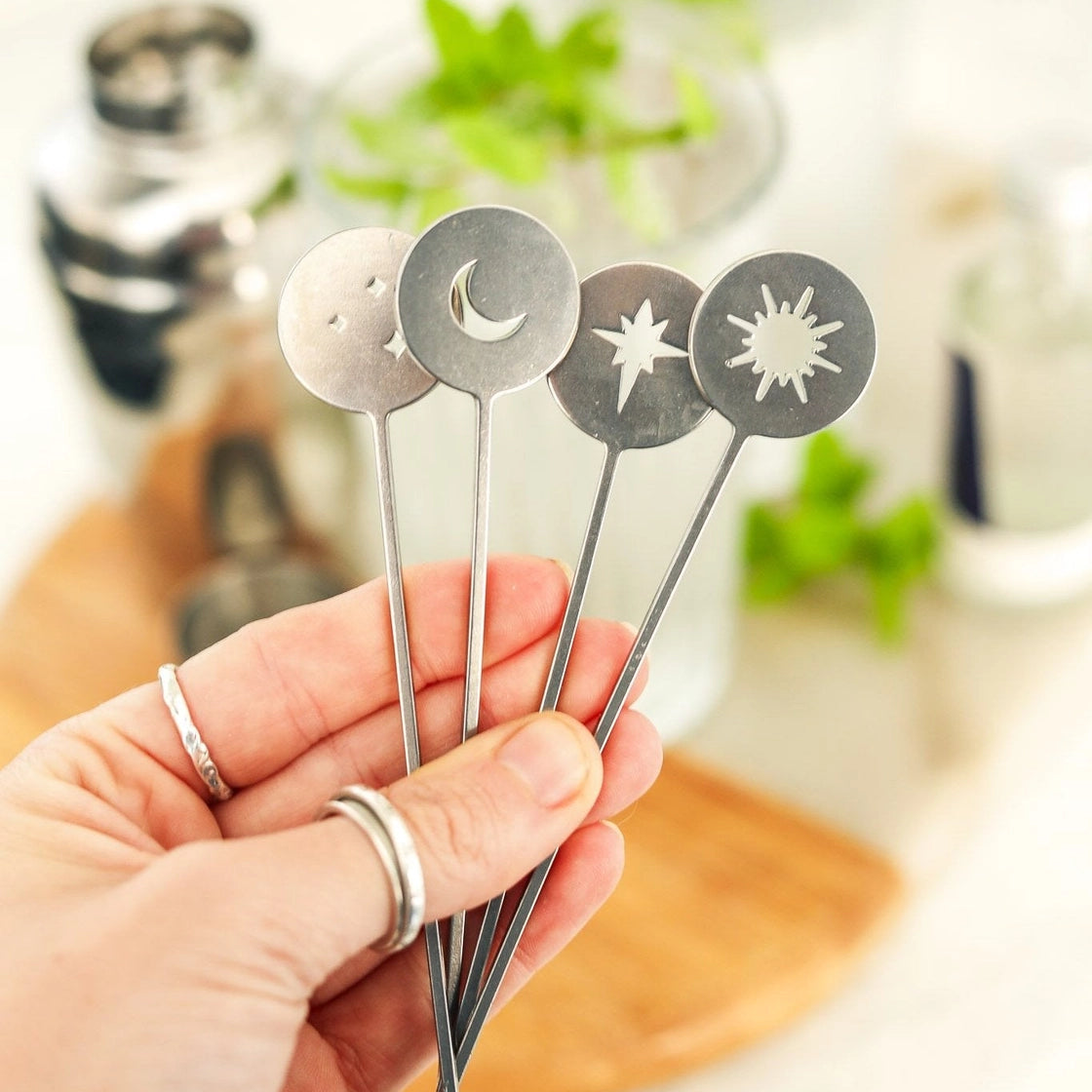 A hand holding four silver metal cocktail stirrers with circular cutout designs: a crescent moon, a star, and two sunburst shapes, against a softly blurred kitchen background.