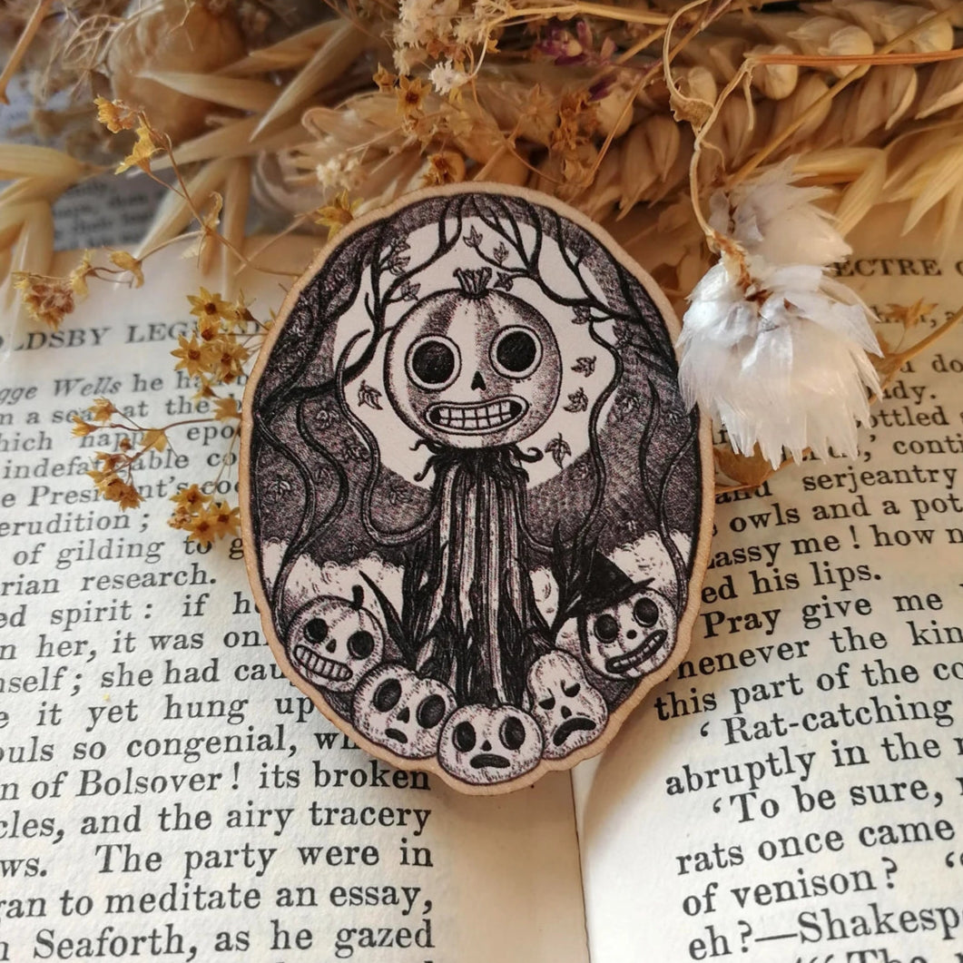 Wooden pin brooch with a decorative black and white, jack'o'lantern design, featuring pumpkins, vines and moonlight. Sat on an open book.