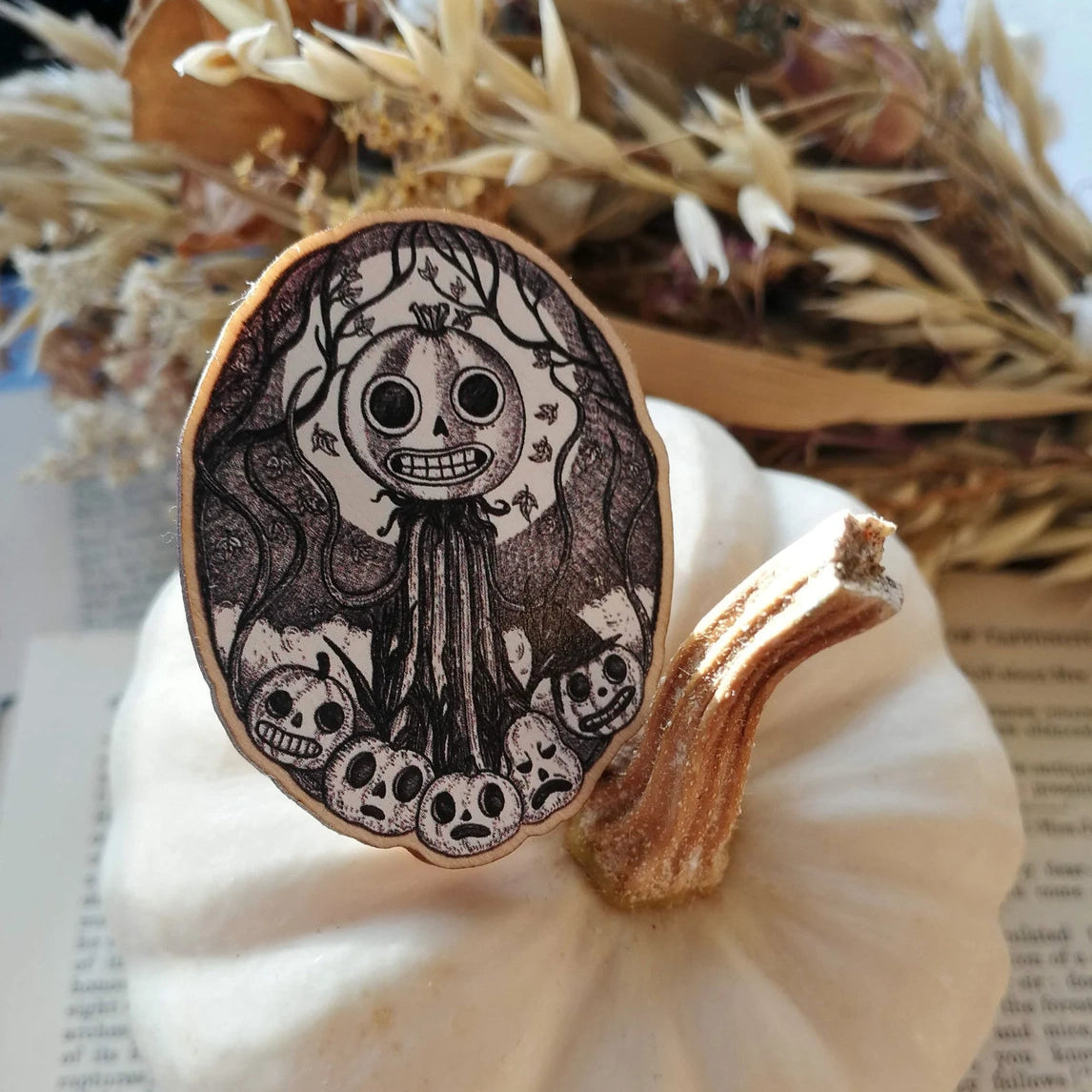 Wooden pin brooch with a decorative black and white, jack'o'lantern design, featuring pumpkins, vines and moonlight. Sat on a white pumpkin.