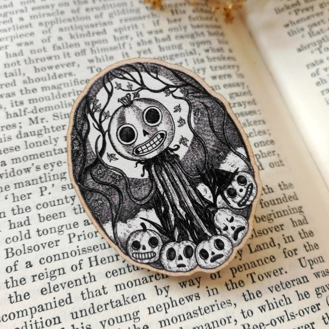 Black and white skull and pumpkin design on an oval brooch placed on an open book with dried flowers.