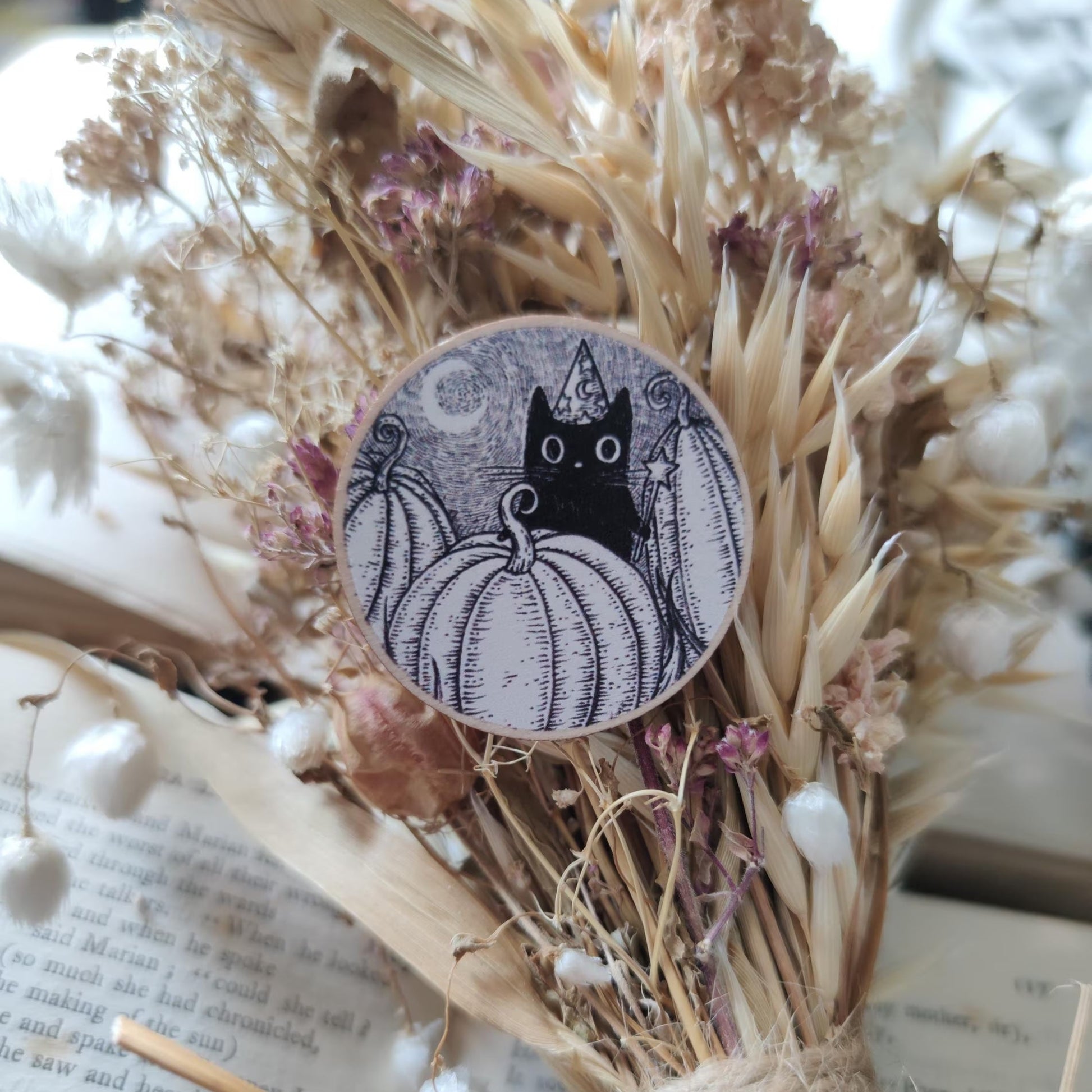Button pin badge with a black cat and pumpkin design on dried flowers and an open book.