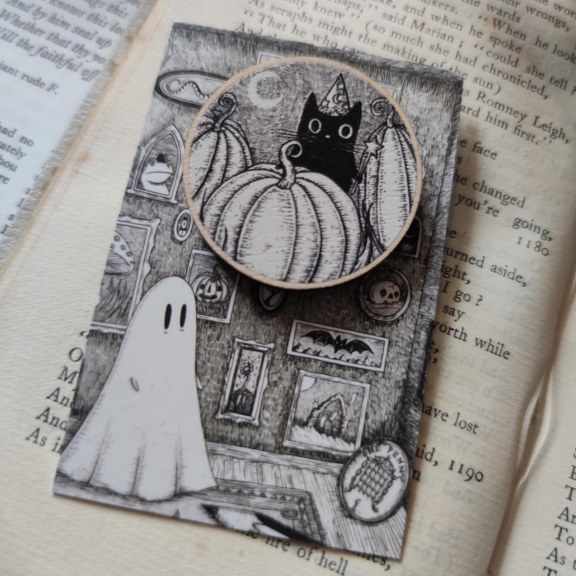 Halloween-themed illustrated woonden pin badge / brooch with a cat, pumpkin, and ghost on an open book.