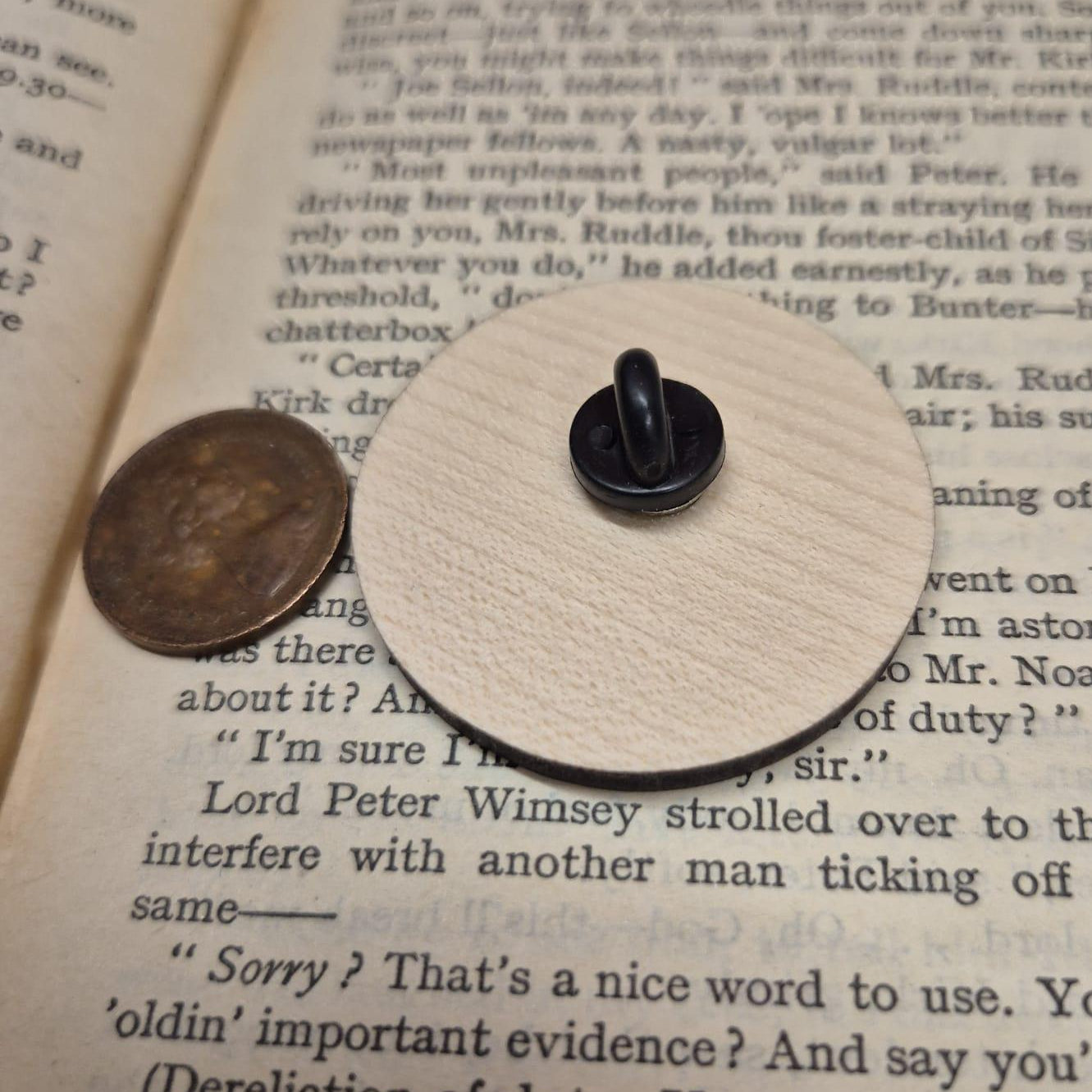 The reverse side of a beige wooden pin with a black button fastening, sat on an open book with a penny coin next to it for size reference.