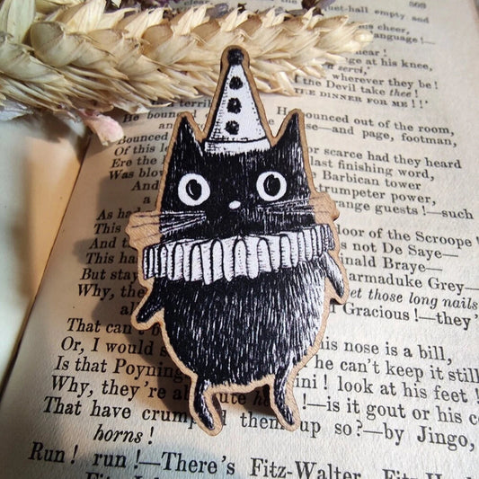 Black and white cat-shaped brooch with a frilly clown collar aand hat, sat on a book page with dried flowers.