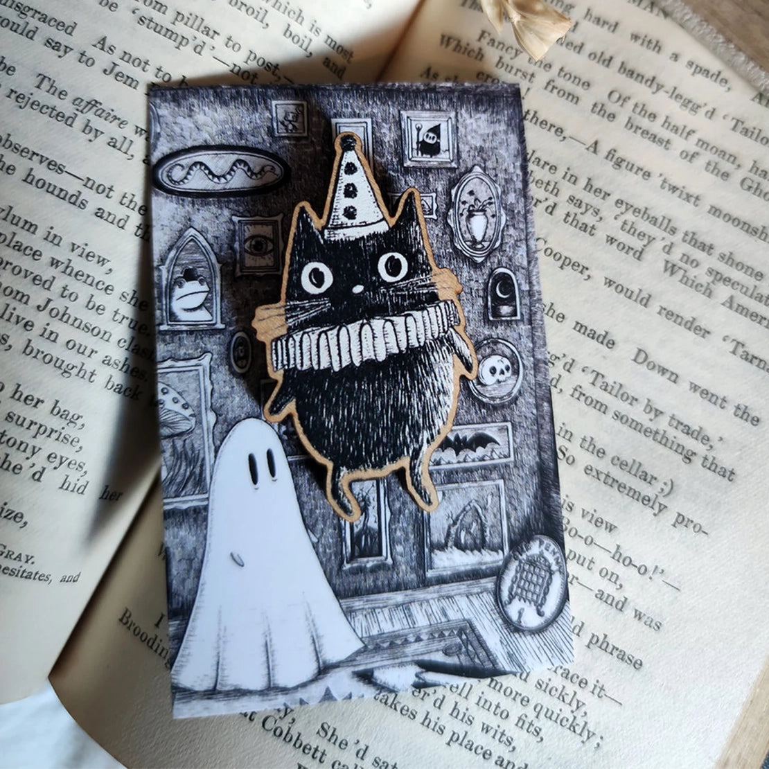 Spooky ghost backing card with a black and white clown cat pin badge attached, sat on an open book.