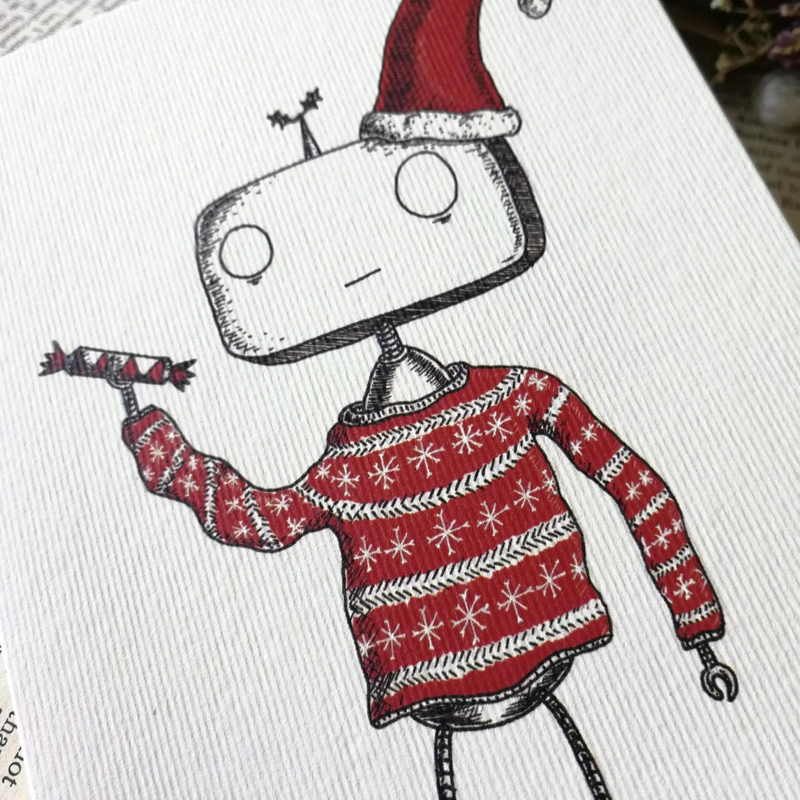Closeup of a white card with illustrated robot wearing a Santa hat and sweater on a book with dried flowers.