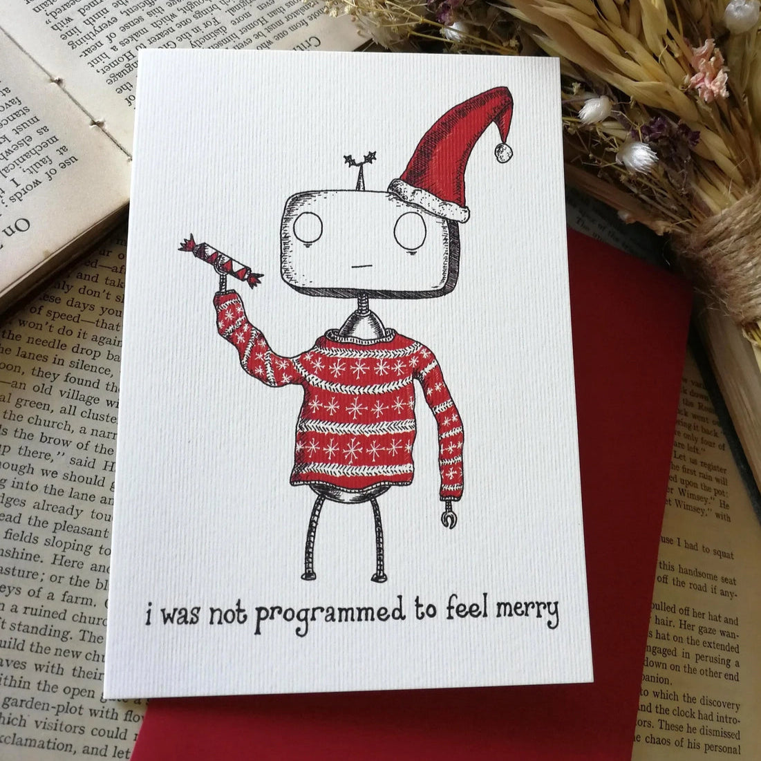 Card with robot wearing a Santa hat and sweater on a book with dried flowers. Includes teh text 'I was not programmed to feel merry'.