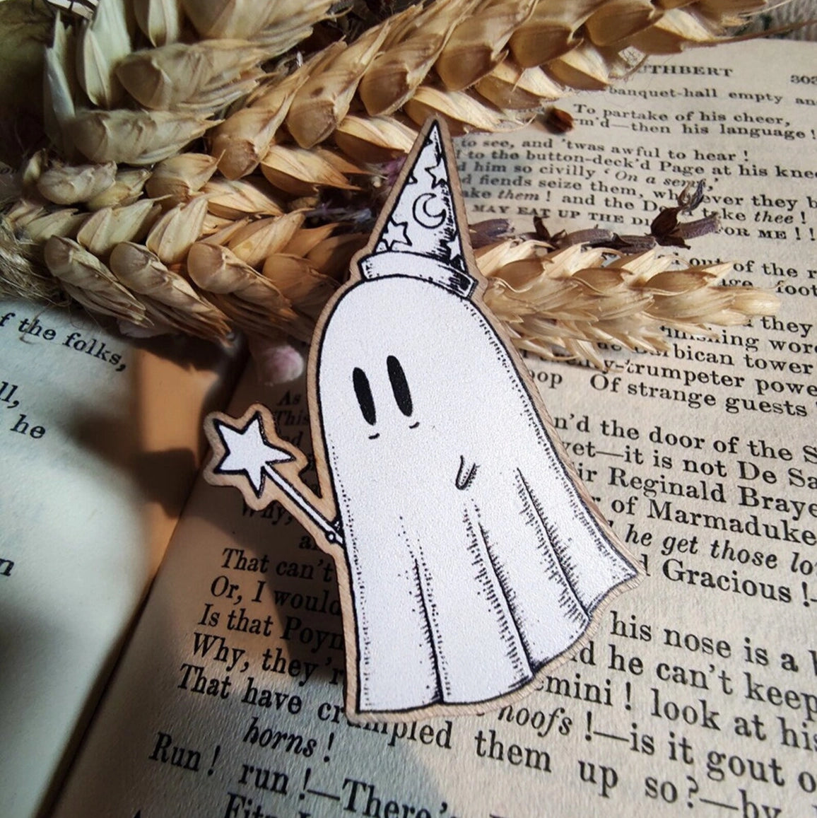 Illustrated black and white ghost with a witch hat and broomstick, sat on an open book with dried plants.