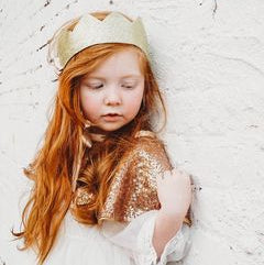 Red haired child modelling the gold crown, leaning on a white wall.