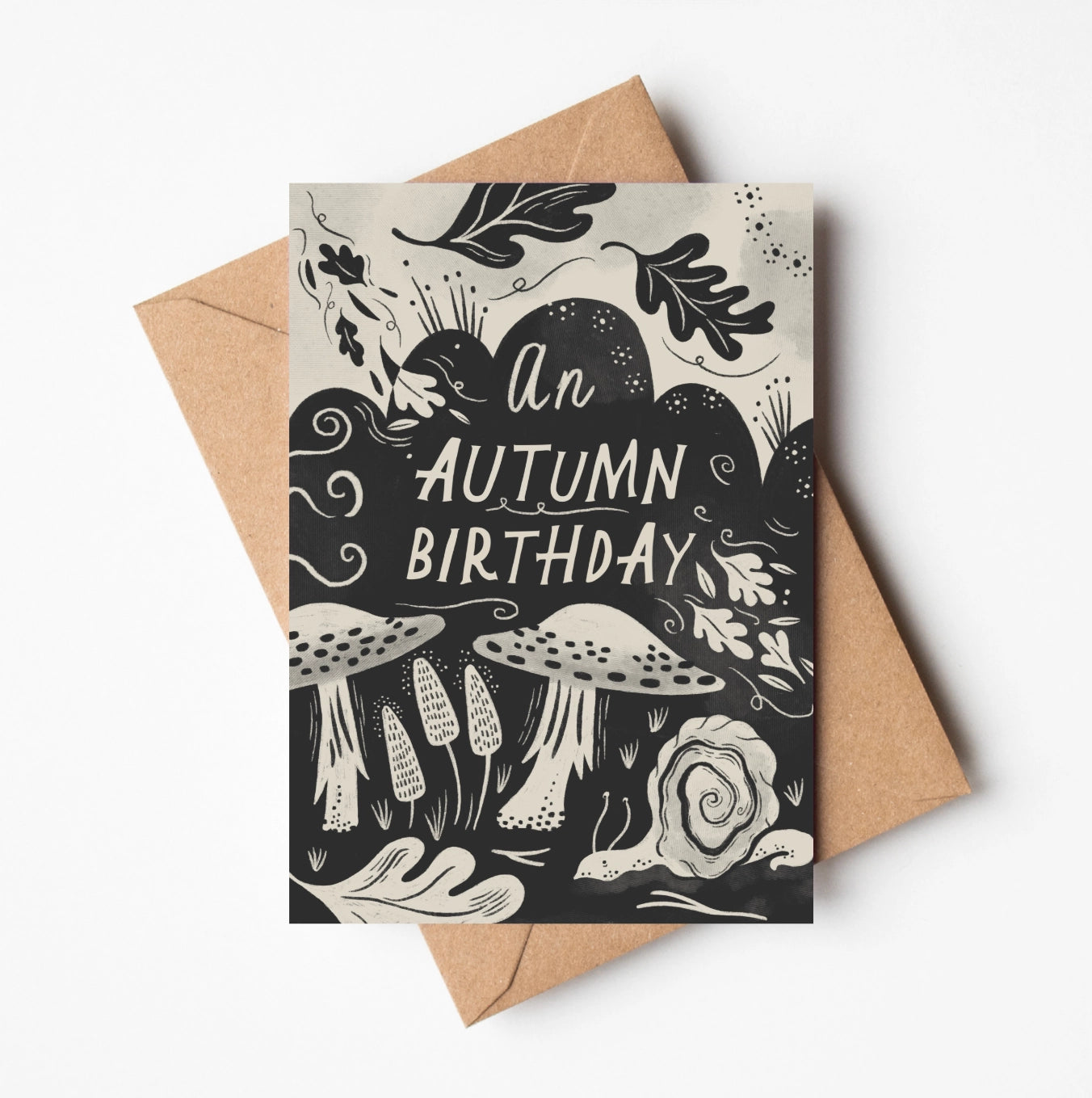Greeting card with 'An Autumn Birthday' text and mushroom illustrations on a black background, with a brown envelope.