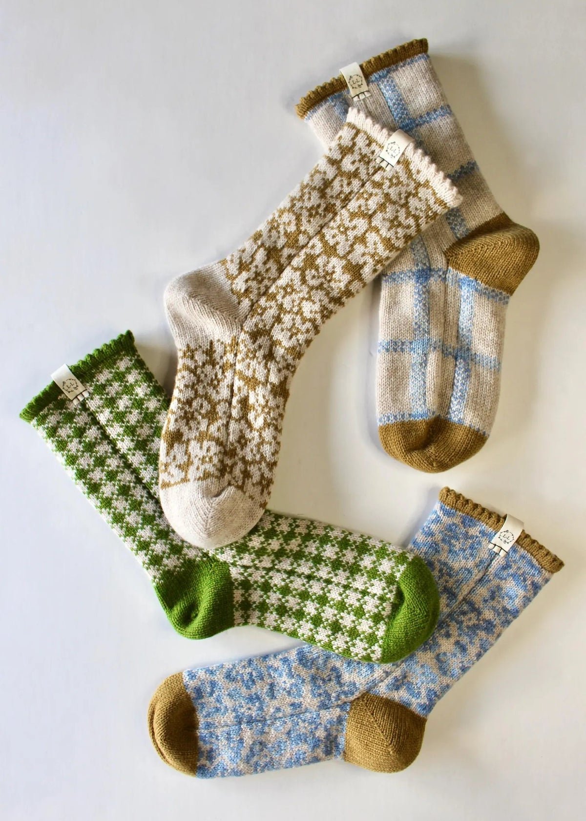 Four pairs of hand-knit socks arranged on a light gray background, featuring different patterns: a green and white houndstooth, a mustard and oatmeal floral, a beige and blue gingham, and a blue and white floral.