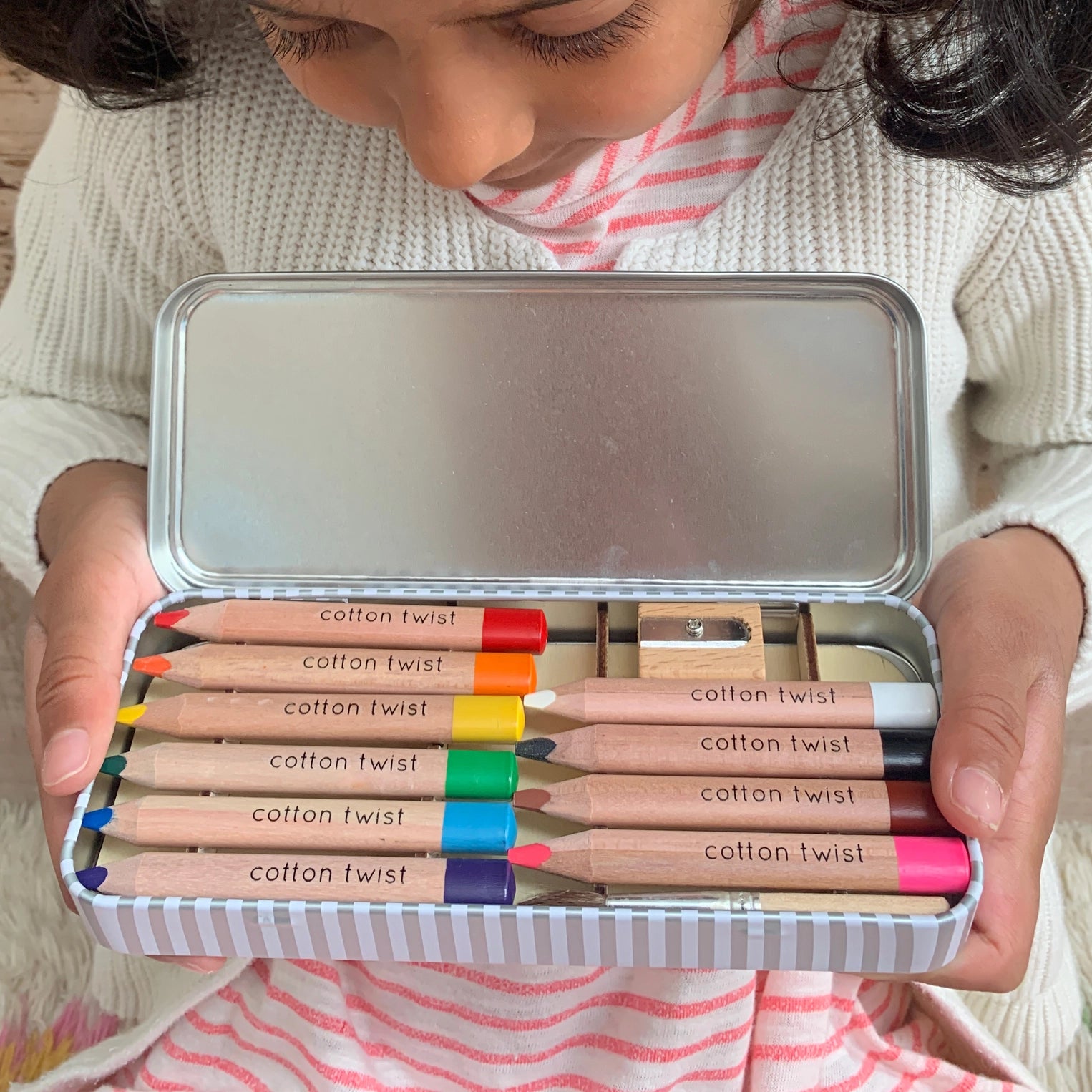 Child holding a set of Cotton Twist colored pencils in a metal tin.