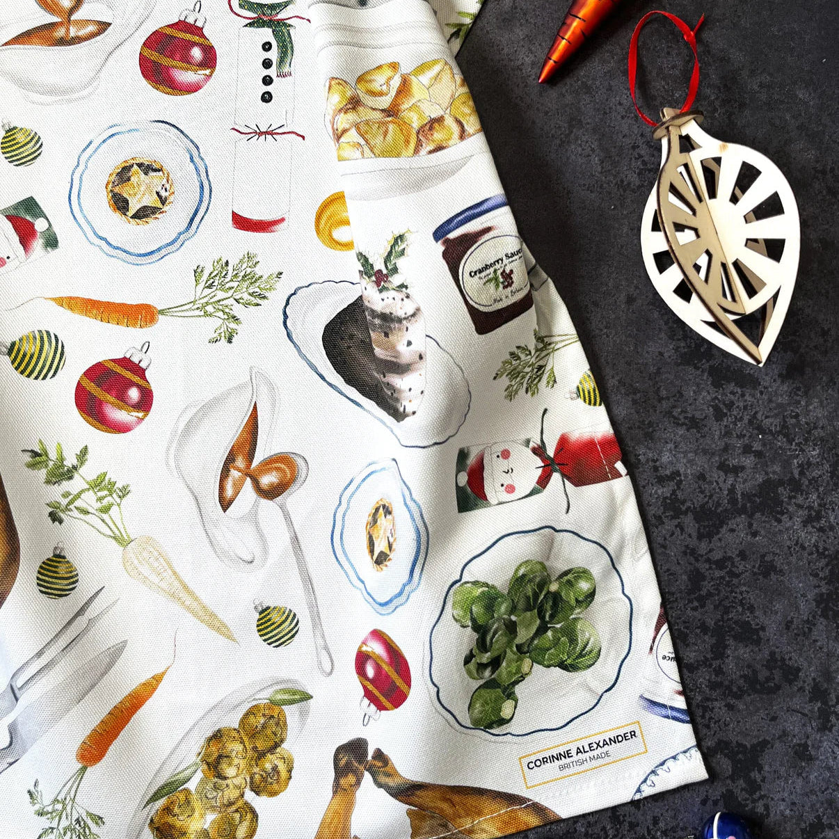 Christmas tea towel with festive food illustrations and a decorative pendant on a dark surface
