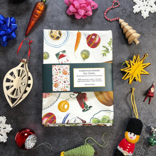 Pack of Christmas-themed tea towels surrounded by decorative ornaments on a dark surface