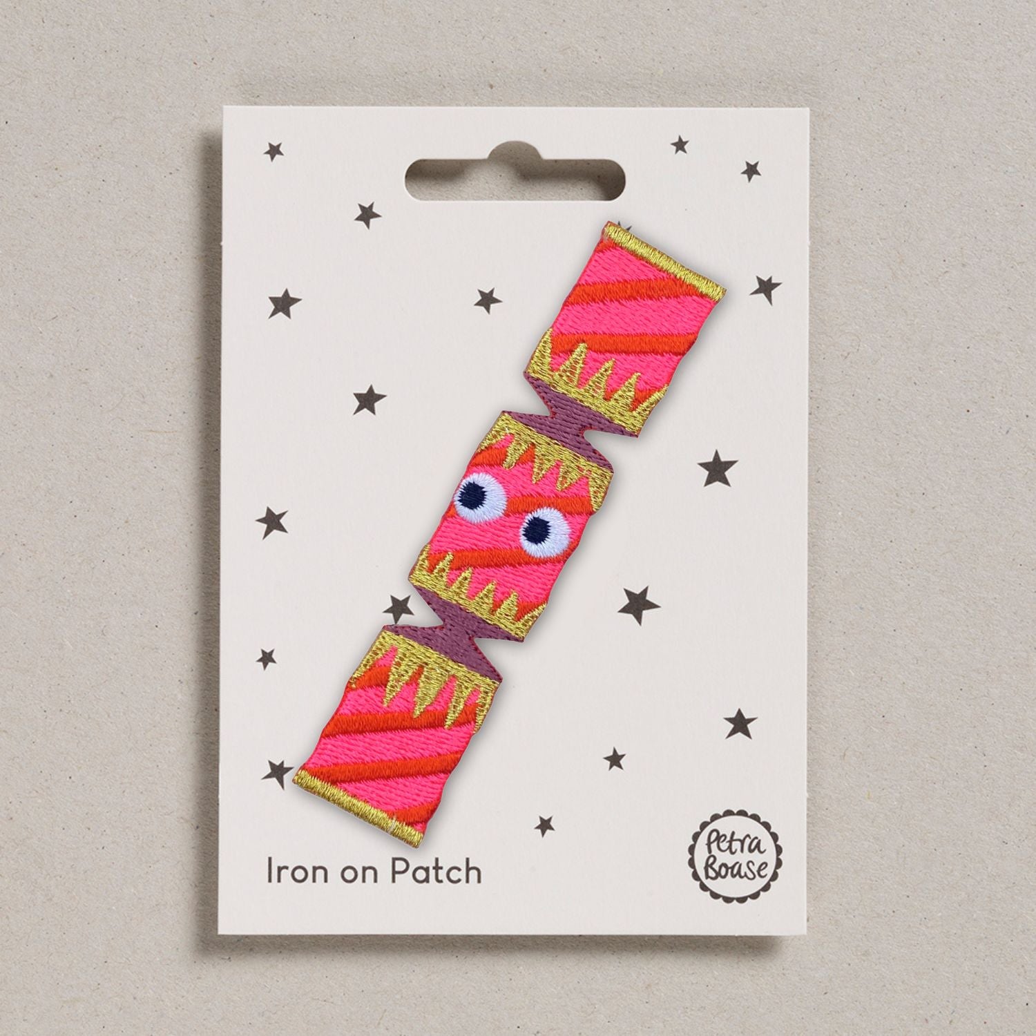 Iron on patch with a pink Christmas cracker design with googly eyes. On a beige background.