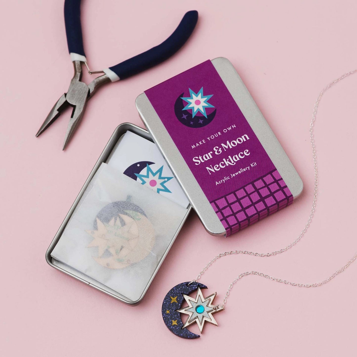 Tin containing DIY necklace pieces, with a finished moon and star necklace on a pink background.