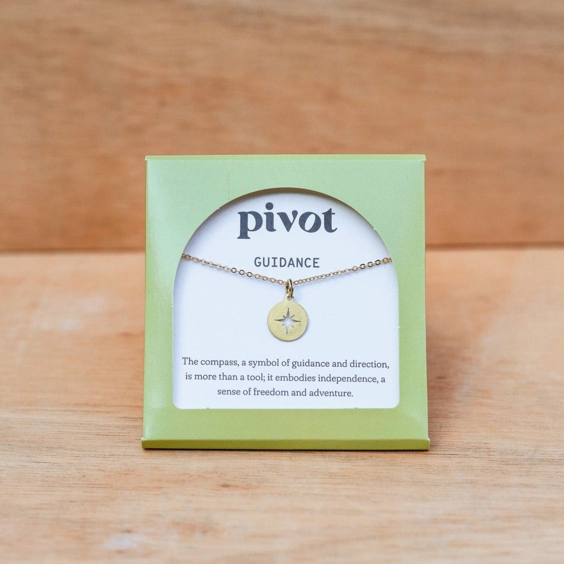 A brass pendant necklace by Pivot, displayed on its sustainable light green packaging card against a wooden background.
