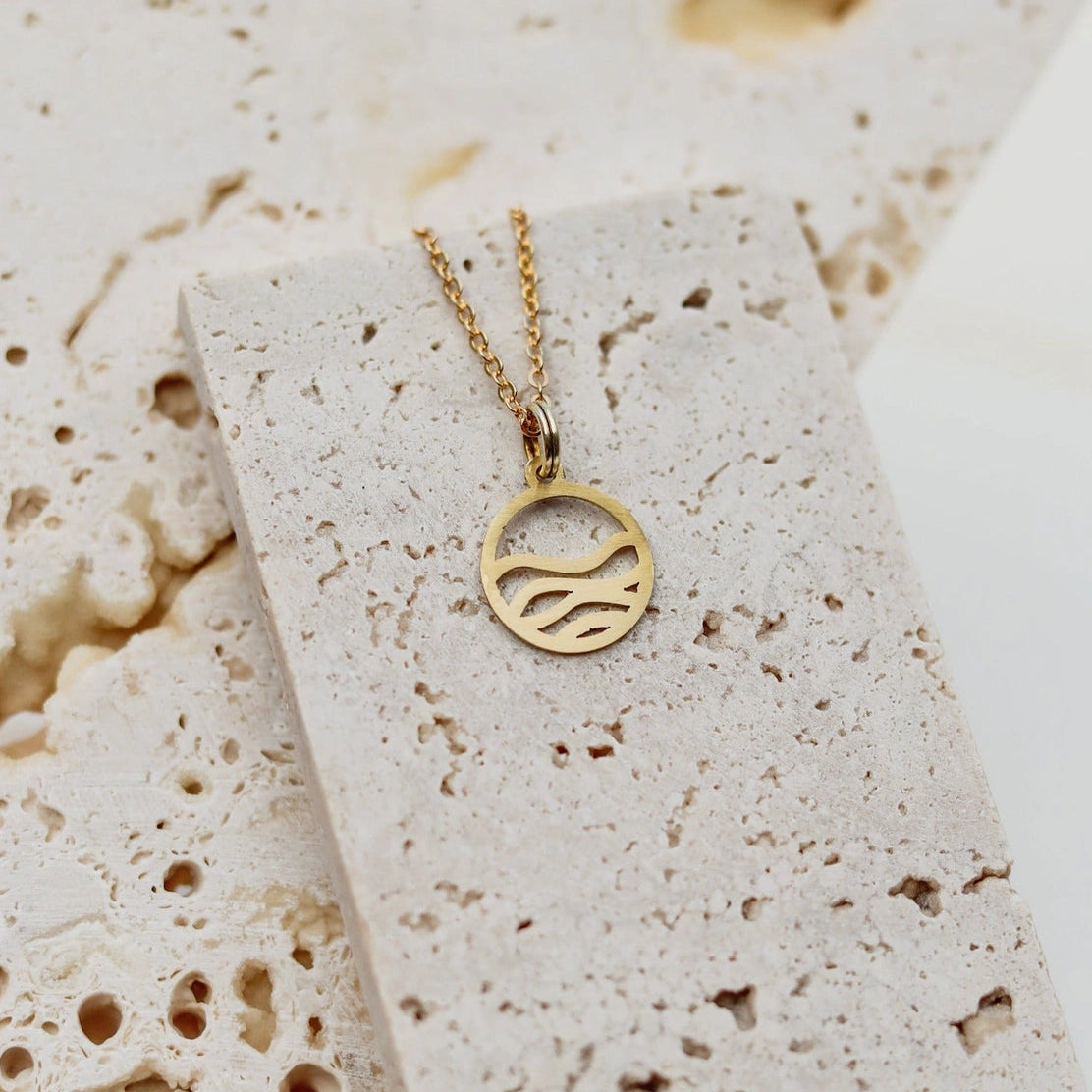A brass pendant necklace with a circular cutout design featuring wavy ocean lines, displayed on a textured beige stone slab.
