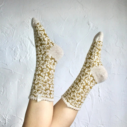 A pair of cream and mustard yellow floral knit socks worn on a person's feet, raised against a textured white wall.
