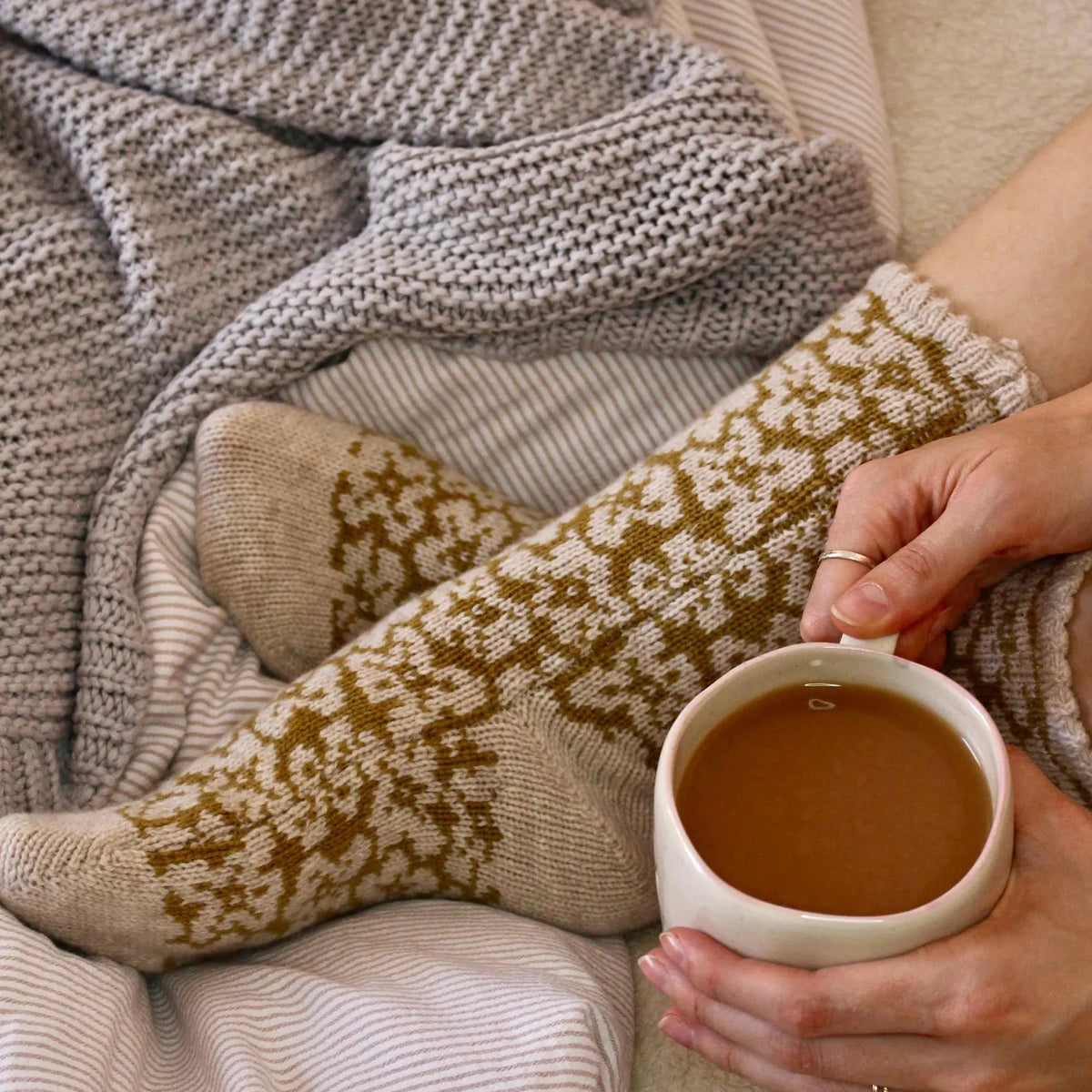 A cozy lifestyle shot of a person's legs wrapped in a gray knit blanket, wearing mustard yellow and oatmeal beige floral pattern knitted bed socks. The person is holding a white ceramic mug filled with a warm brown beverage, set against a background of striped bedding.