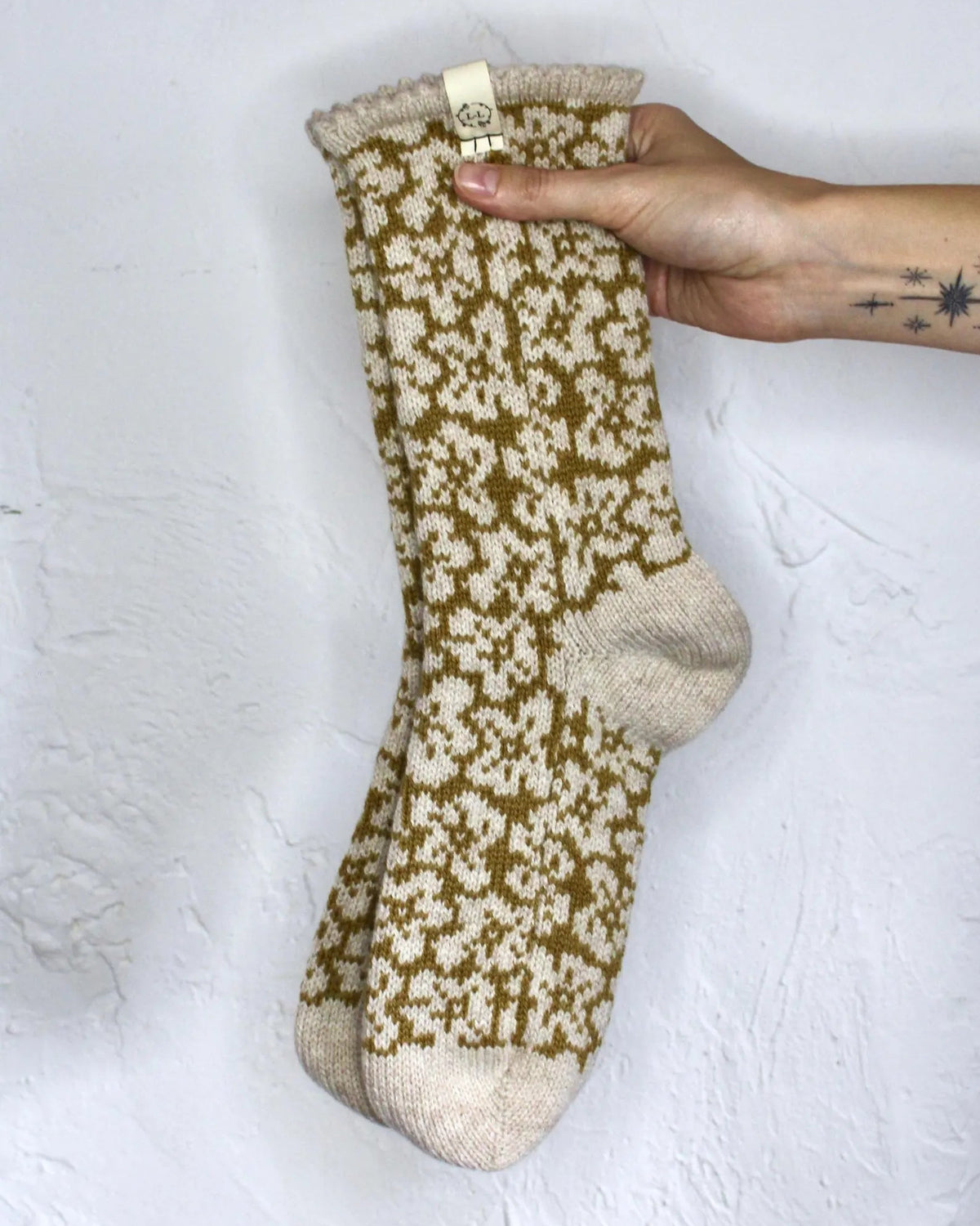 A hand holds up a pair of cream and mustard yellow patterned knit bed socks against a textured white wall.
