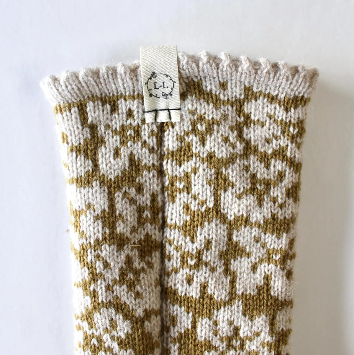 A close-up of a hand-knit sock with a cream and mustard yellow floral pattern, featuring a small white Lizzie Lou Textiles brand tag at the cuff.