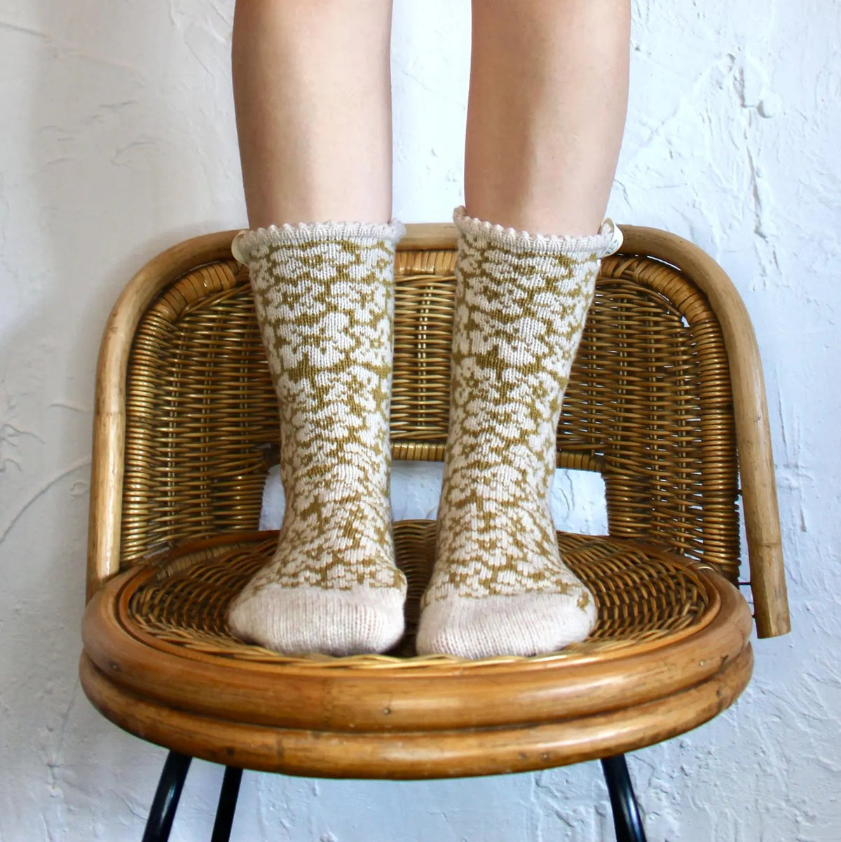 A person's legs are shown wearing mustard yellow and oatmeal beige patterned knit socks while standing on a round wicker chair against a white textured wall.