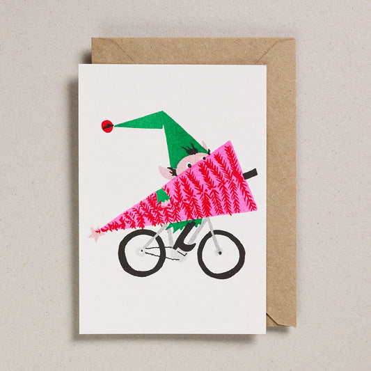 Greeting card with an illustrated elf riding a bike carrying a pink Christmas tree,on a white background with a brown envelope.