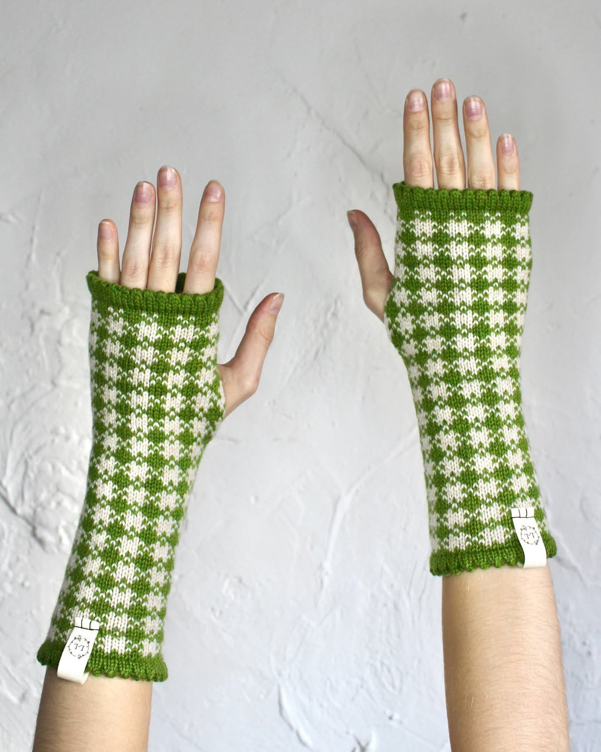A pair of green and cream gingham knit fingerless gloves worn on hands, shown against a textured light gray background.
