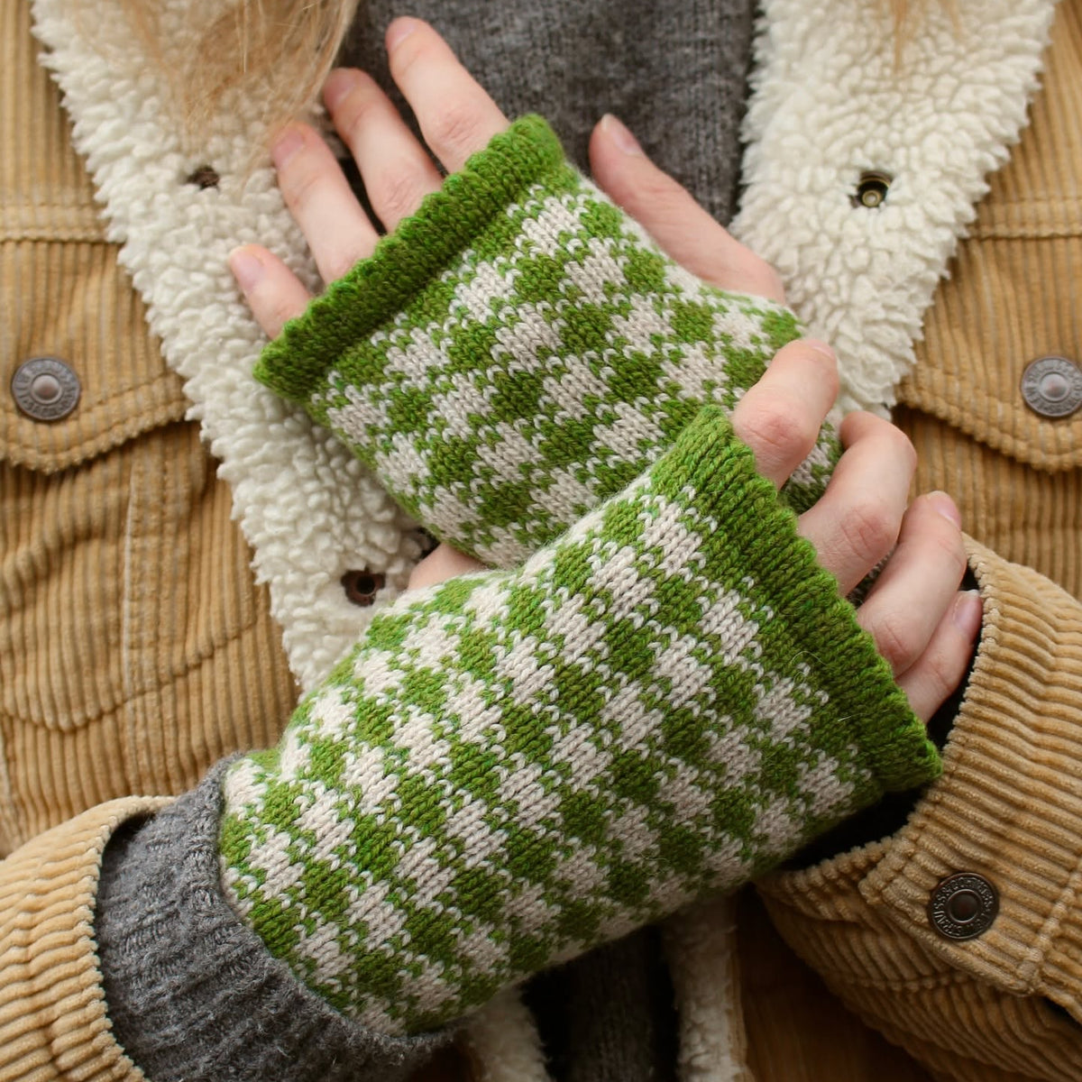 Green and white checkered knit fingerless wrist warmers worn on a person's hands, shown against a brown corduroy jacket with a cream sherpa lining.