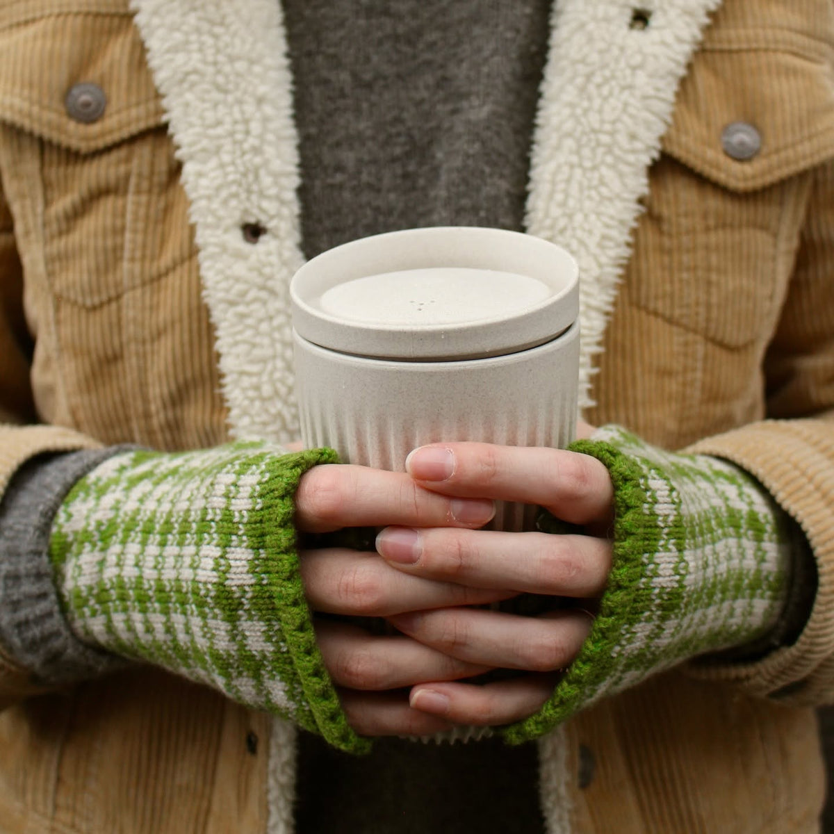 A person wearing a tan corduroy jacket with a cream sherpa lining, a gray sweater, and green-and-white check knit fingerless gloves, holding a white coffee cup.