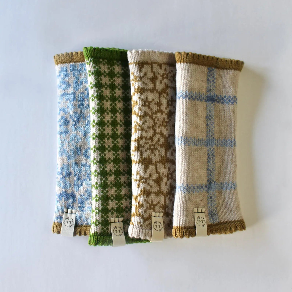 Four knitted lambswool fingerless gloves / wrist warmers,  with different knit patterns in blue, green, tan, and cream, arranged side-by-side on a plain white background.