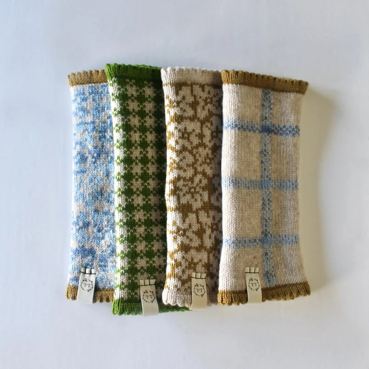 Four knitted fingerless gloves with different knit patterns in blue, green, dijon yellow, and oatmeal, arranged side-by-side on a plain white background.