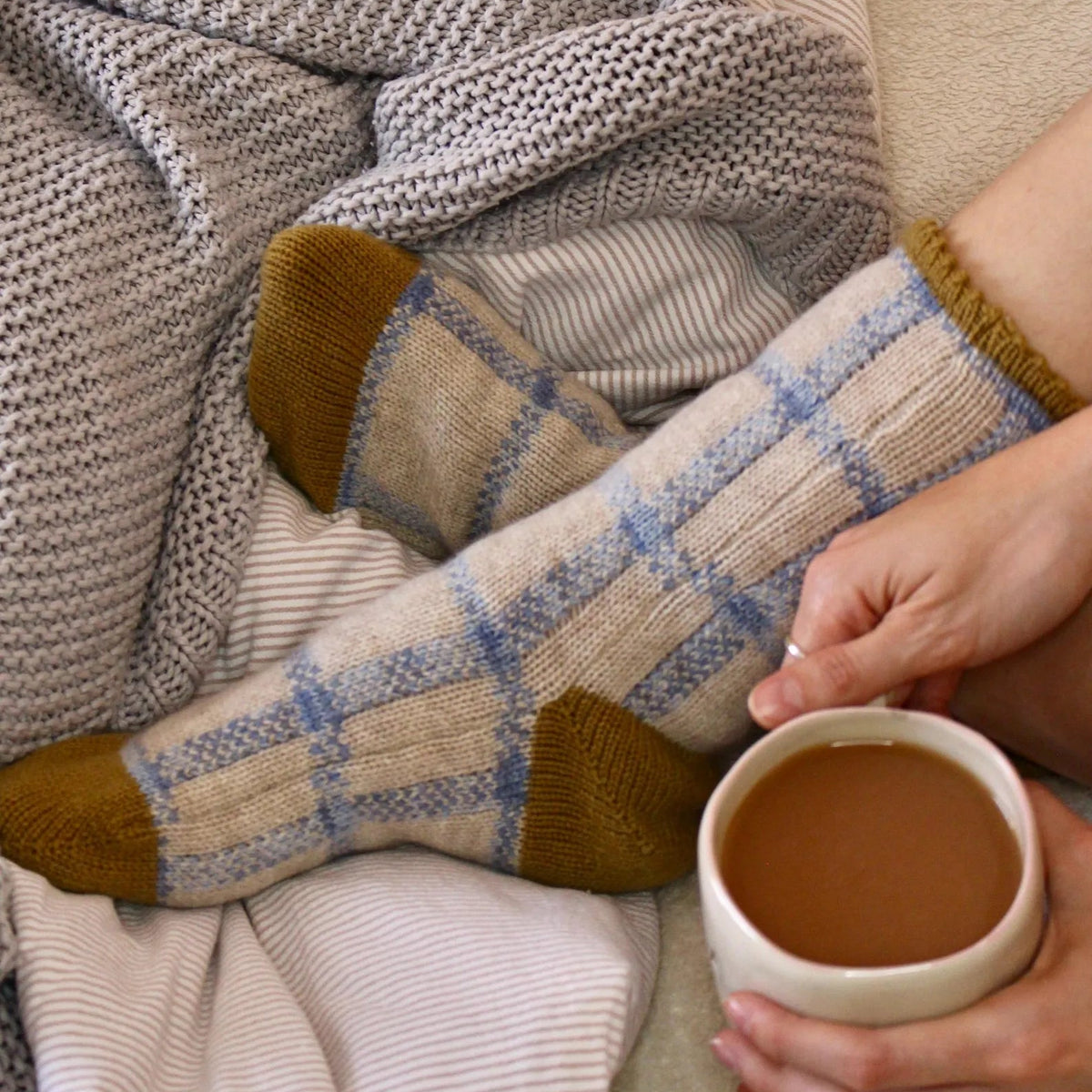 A close-up lifestyle shot of a person's feet wearing cozy knit socks with a beige and blue plaid pattern and mustard yellow toes, wrapped in a striped blanket and a chunky gray knit throw, while holding a cup of coffee.