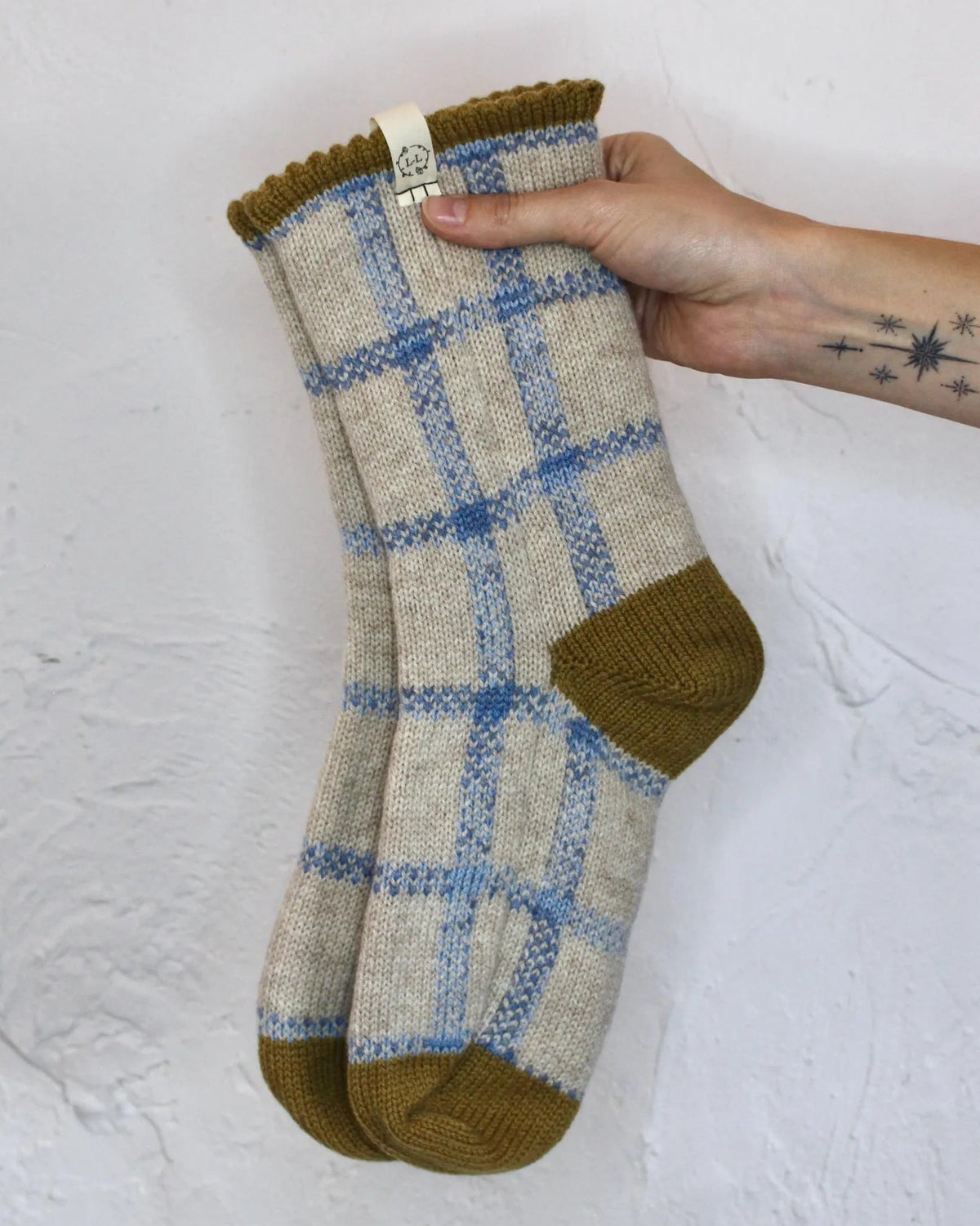 A pair of knit bed socks held up by a hand against a white textured wall. The socks have an oatmeal crea, base with a blue plaid pattern and mustard yellow accents on the heels, toes, and cuffs.