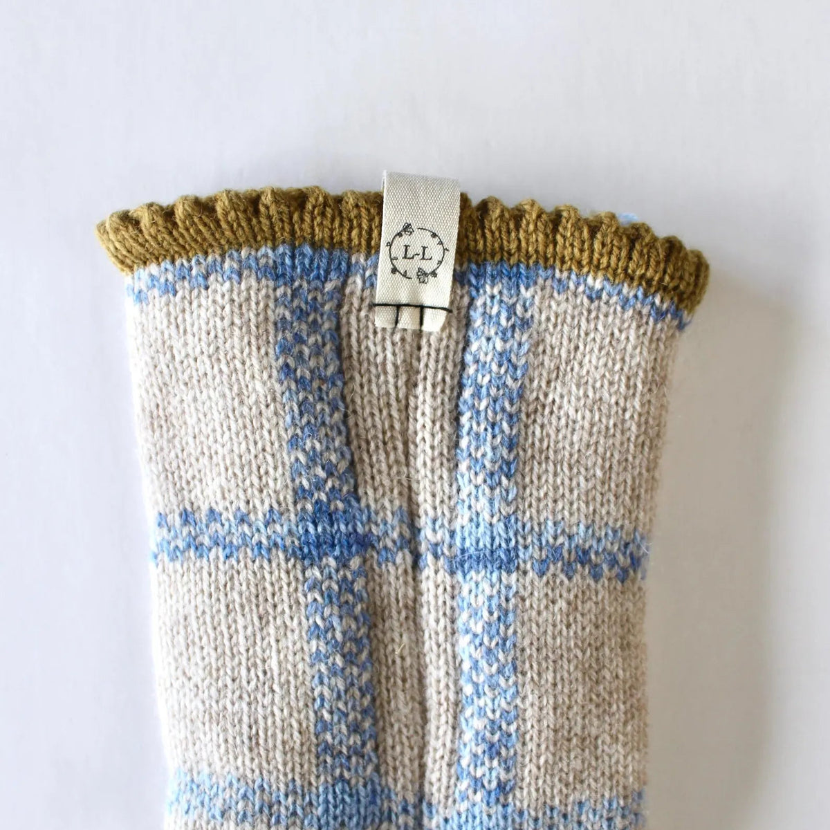 A close-up of a hand-knit bed sock featuring a beige and light blue plaid pattern with a mustard yellow ribbed cuff with a Lizzie Lou brand label, shown against a plain white background.