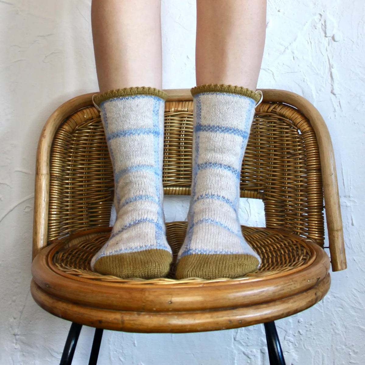 A person's legs are shown wearing blue and cream gingham socks while standing on a wicker chair against a white textured wall.
