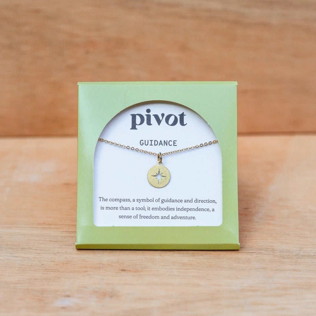 A brass compass pendant necklace by Pivot, displayed on its light green packaging card against a wooden background.