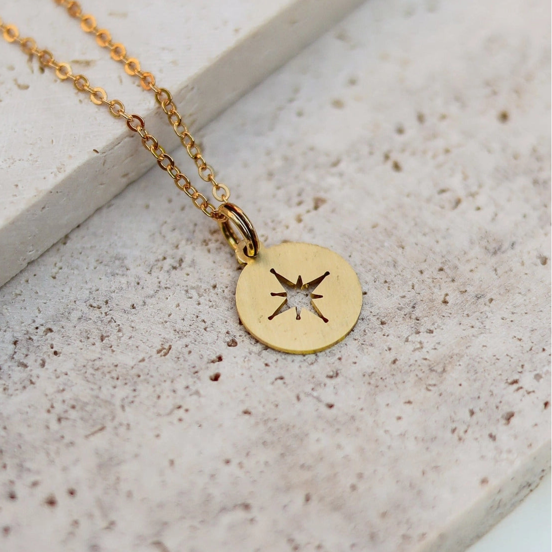 A brass chain necklace with a small round gold disc pendant featuring a cutout star design, resting on a textured light beige stone surface.
